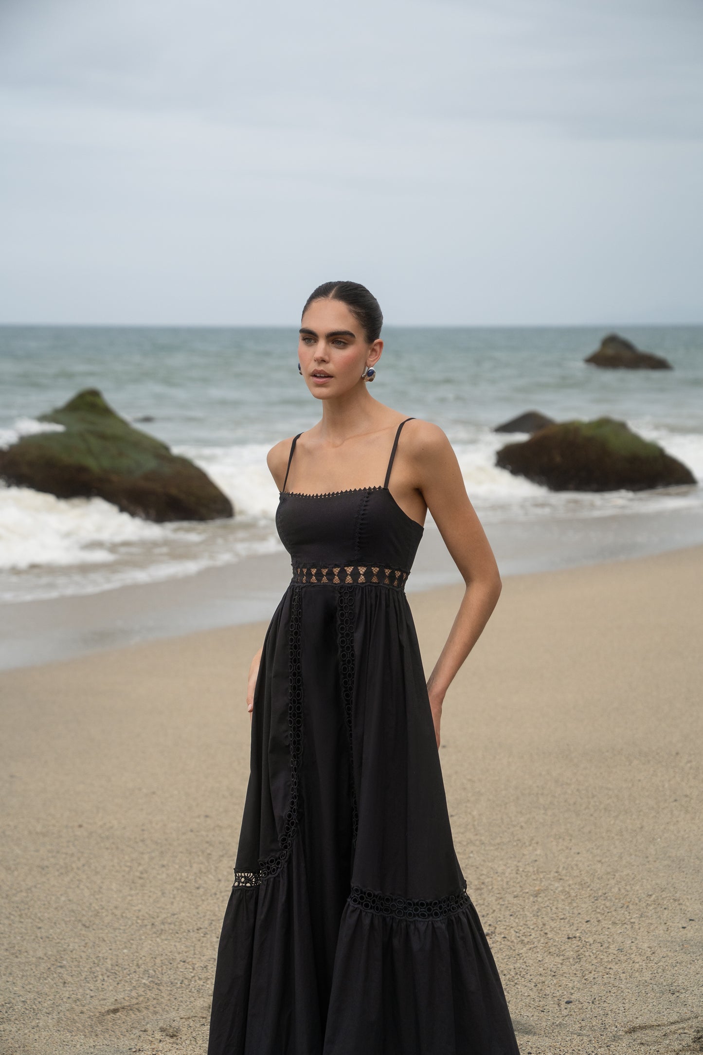 Woman in a black dress standing on a beach with ocean waves and rocks in the background