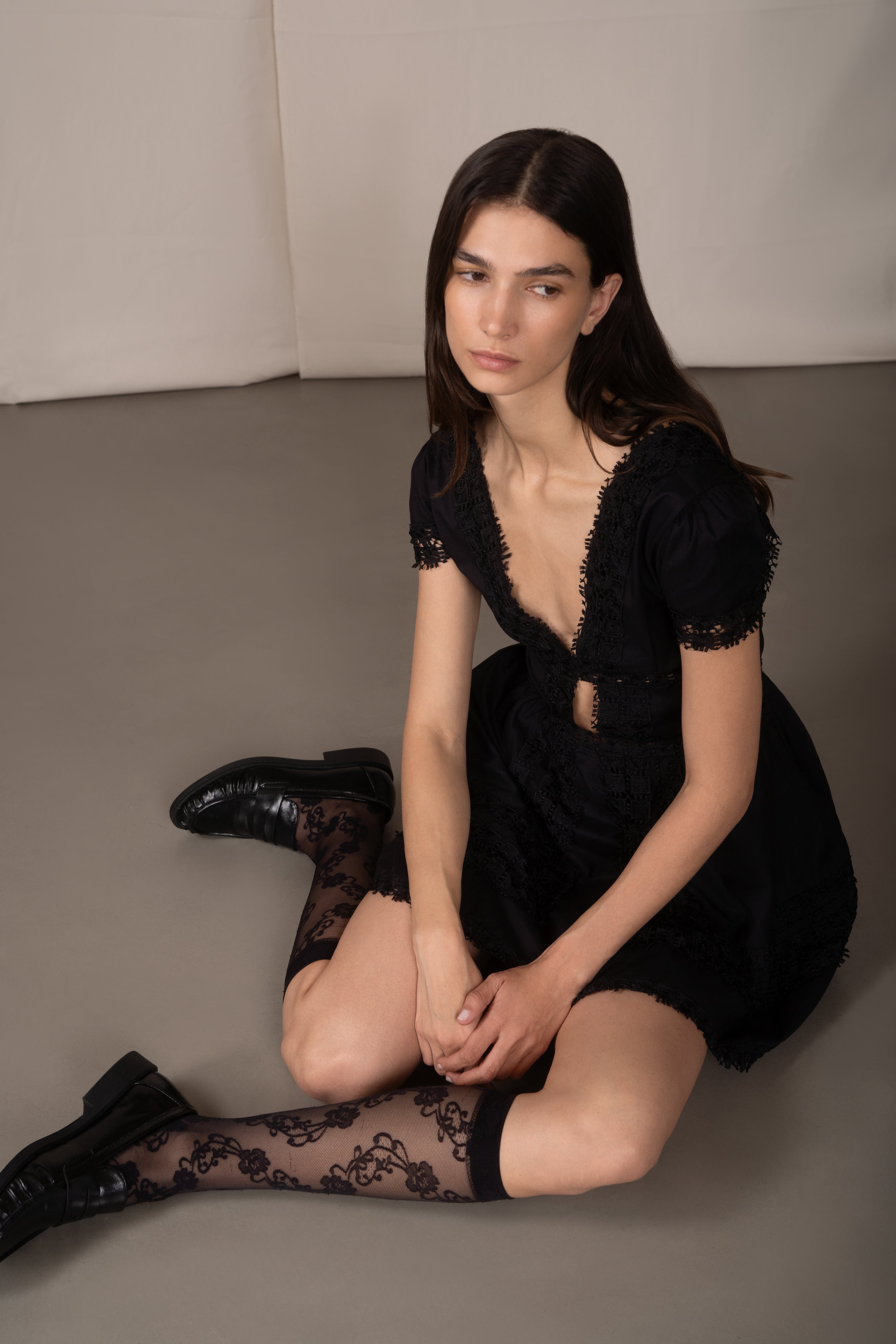 Woman wearing a black dress and stockings sitting on a gray floor.