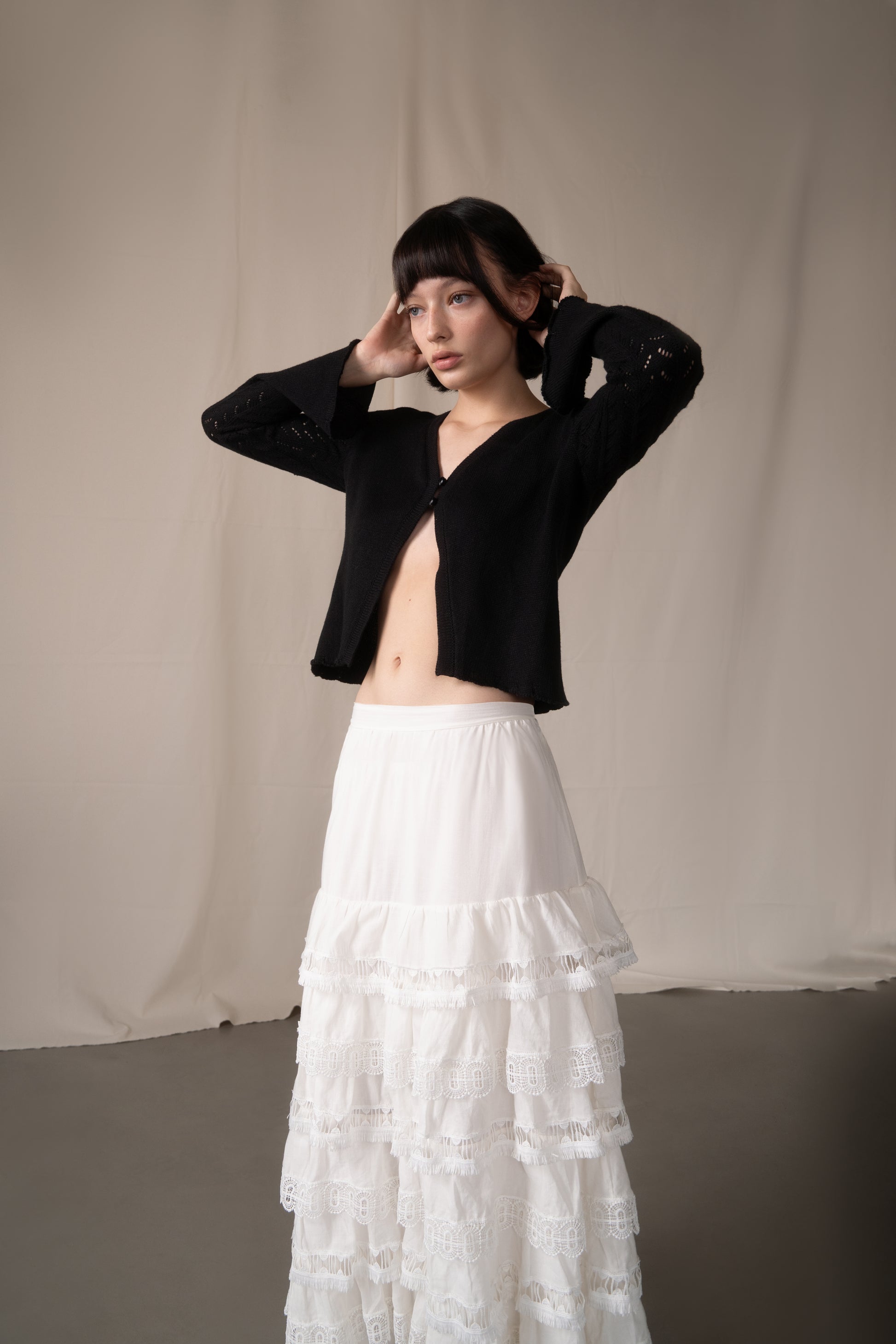 Woman wearing a black cropped jacket and white ruffled skirt against a beige curtain background