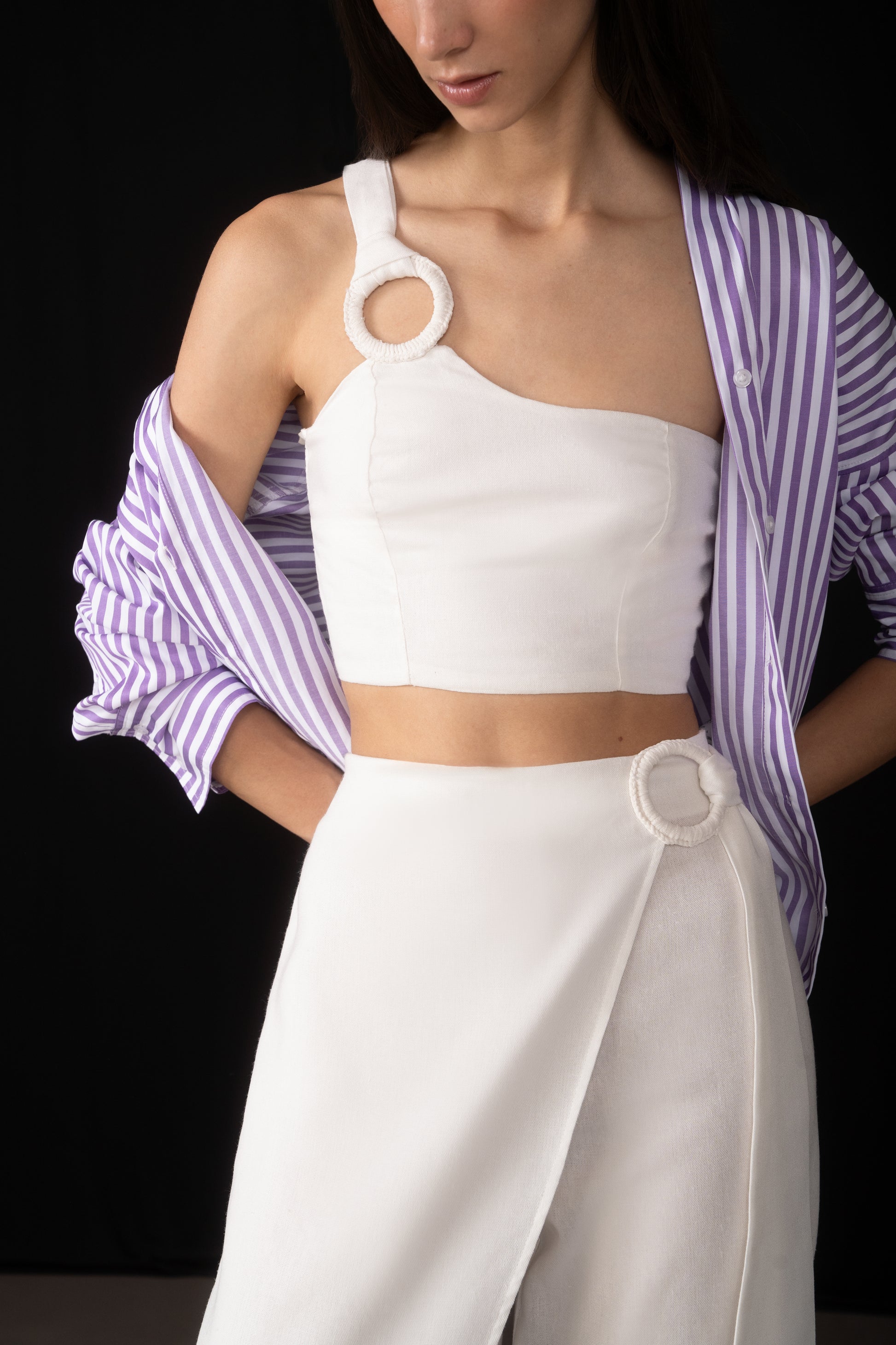 Woman wearing a white top and pants with a purple striped shirt over her shoulders against a black background
